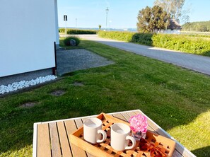 Ein Holztisch mit zwei weißen Tassen und einer rosa Blume, auf einer Rasenfläche mit einem weißen Gebäude und einer Straße im Hintergrund.