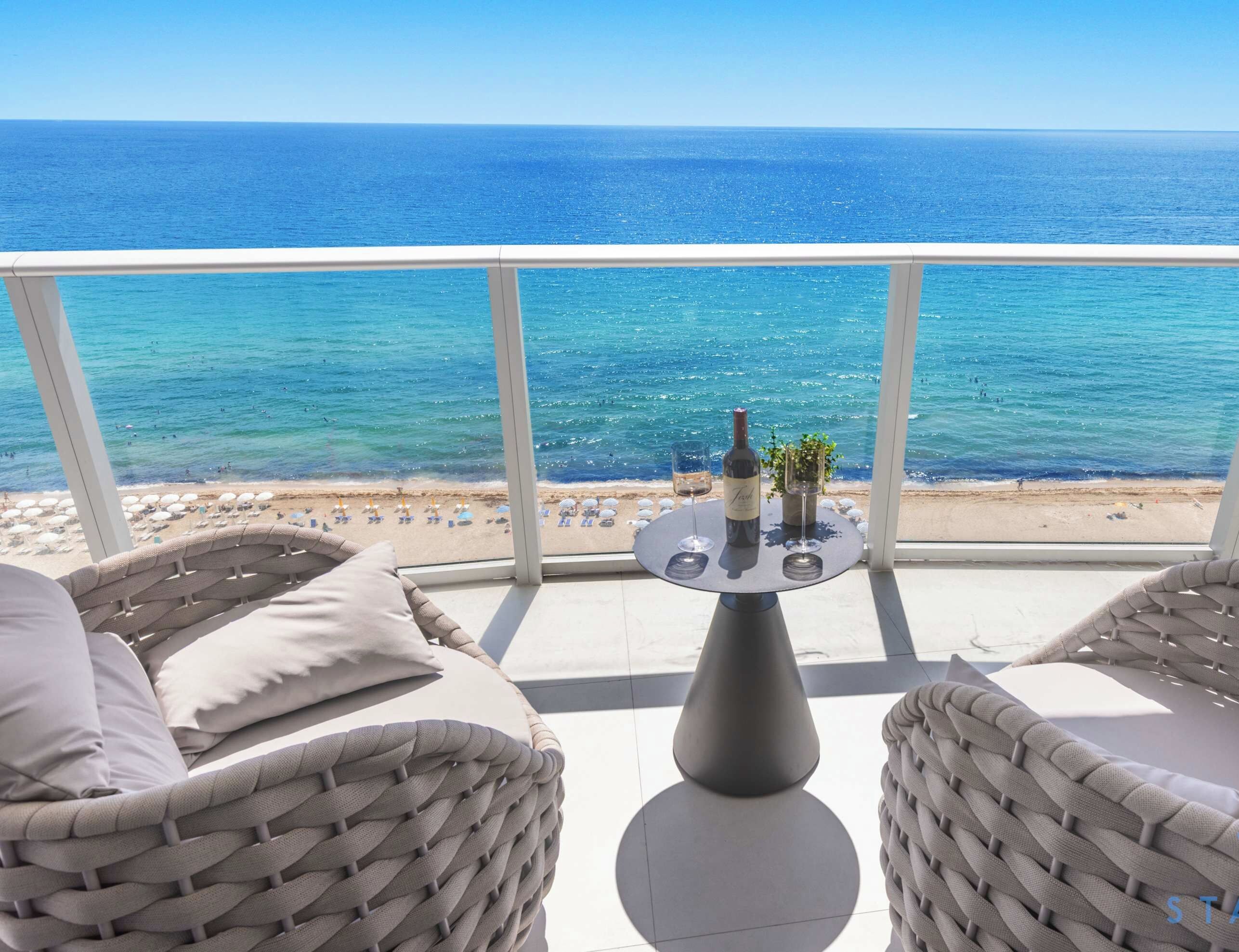 A balcony with wicker furniture, a table with a bottle and glasses, and a view of the sea and beach.