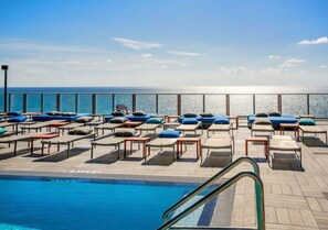 A poolside area with lounge chairs and tables, a pool, and a view of the ocean.