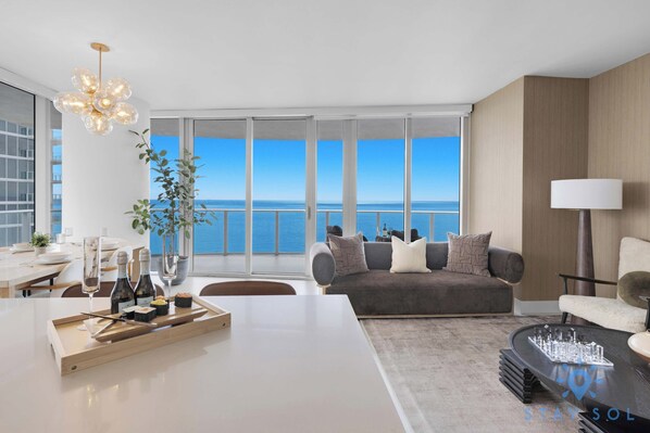 A modern living room with a large window, a dining area, and a seating area with a view of the sea.