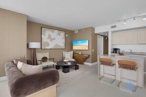 A modern living room with a sofa, a coffee table, and a TV. There is a kitchen area with a bar and stools.
