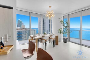 A modern dining area with a large table set for six, large windows offering a view of the sea and cityscape, and a potted plant on the right.