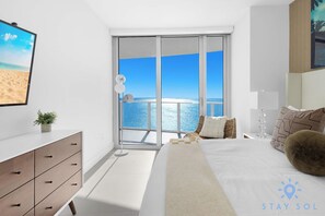 A modern bedroom with a large bed, a nightstand, a dresser, and a view of the ocean through a sliding glass door.