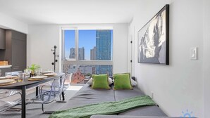 Un apartamento moderno con zona de comedor, sofá cama, ventana grande con vista a la ciudad y un cuadro en la pared.