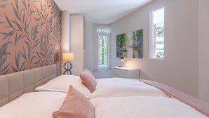 Een slaapkamer met een groot bed, een nachtkastje met een lamp, een raam met uitzicht op bomen en een muur met een bladmotief.