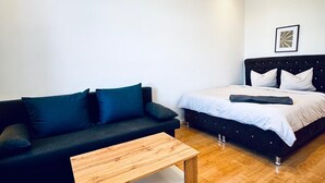 A modern room with a wooden coffee table, a black sofa with blue cushions, and a bed with white bedding.