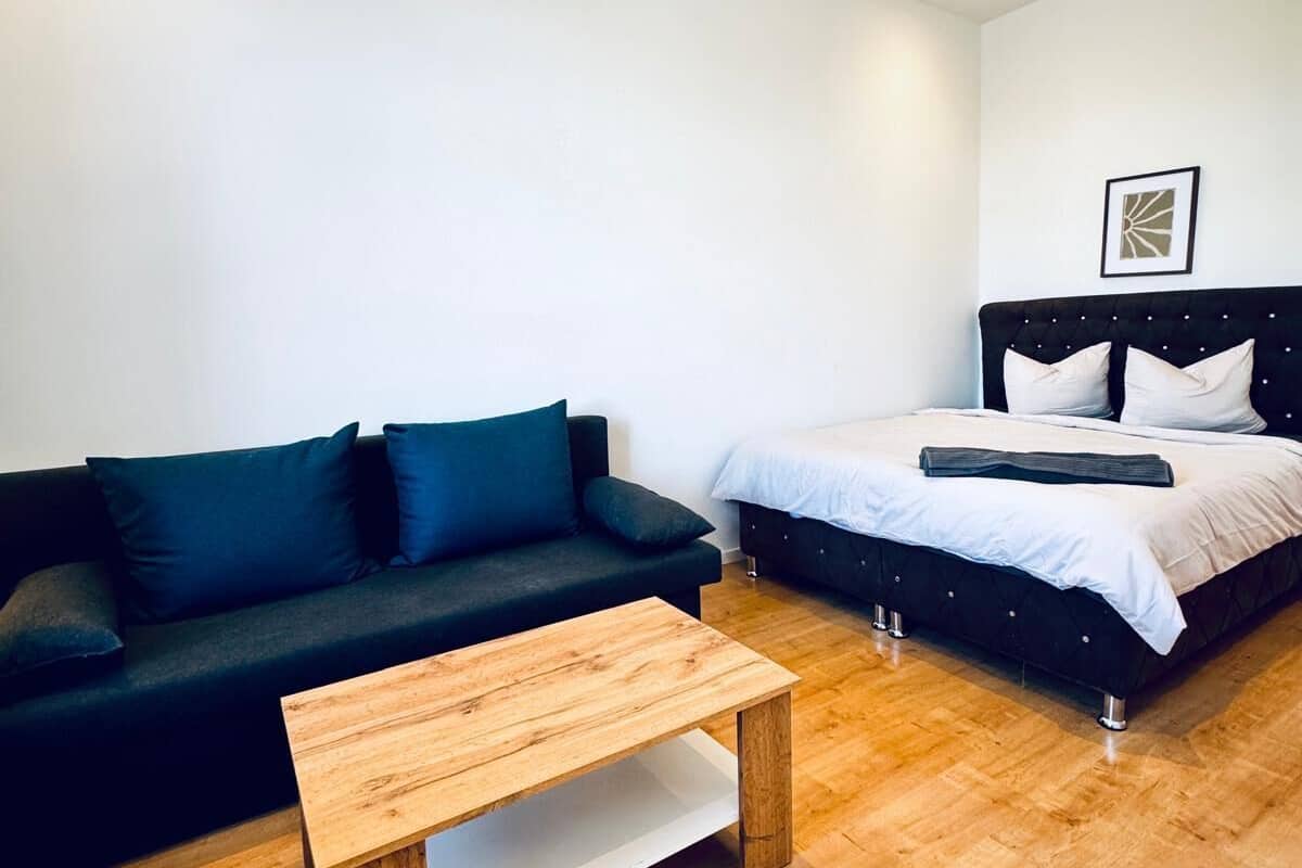 A modern room with a wooden coffee table, a black sofa with blue cushions, and a bed with white bedding.