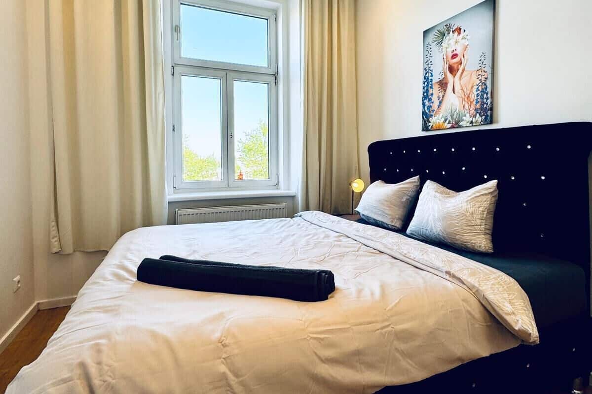 A bedroom with a large bed, a window with curtains, a wall-mounted picture, and a bedside lamp.