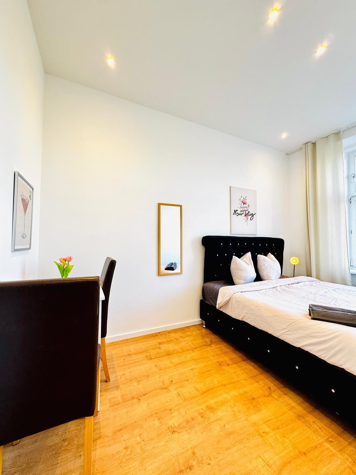 Une chambre moderne avec un lit recouvert de tissu noir, du linge de lit blanc, un plancher en bois, un bureau avec une chaise et un tableau encadré au mur.