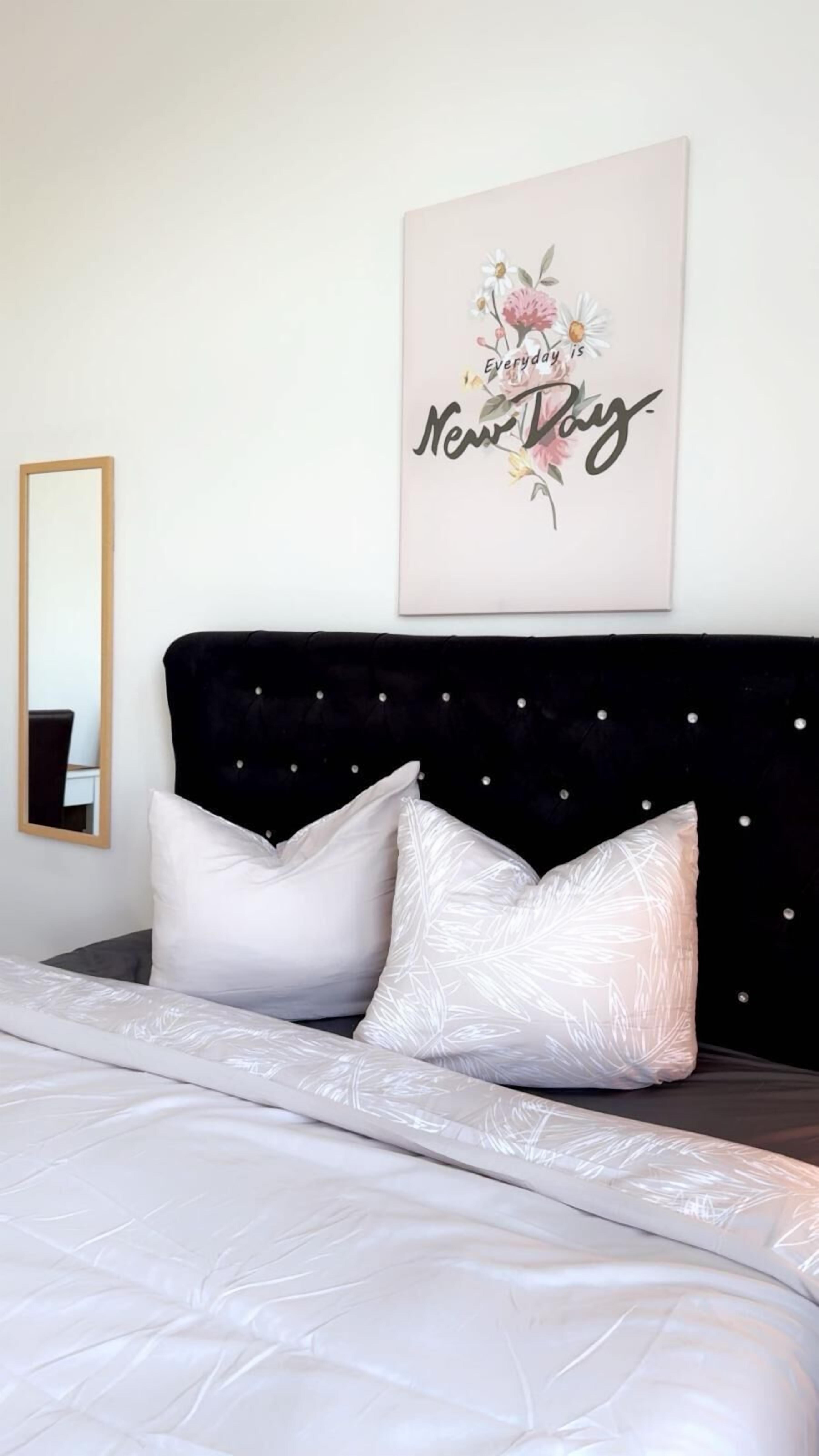 Een net opgemaakt bed met witte beddengoed en twee decoratieve kussens, een zwart bekleed hoofdbord met kleine knopen, en een spiegel aan de muur.