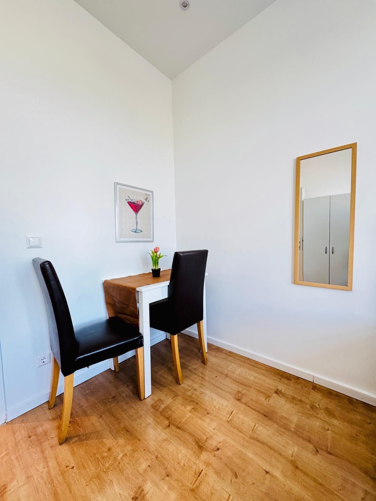 A dining area with a white table, two black chairs, a wooden floor, a framed picture on the wall, and a mirror on the right.
