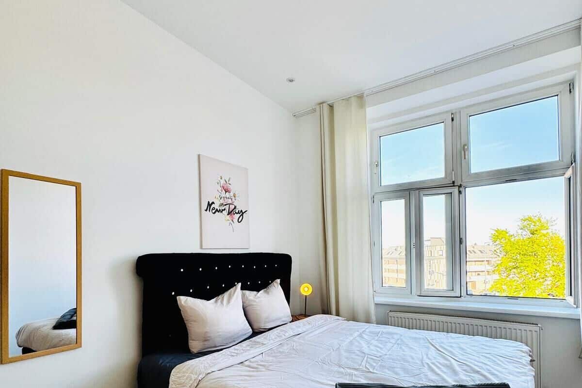A bedroom with a bed, a bedside table, a mirror, and a window with a view of buildings and trees.