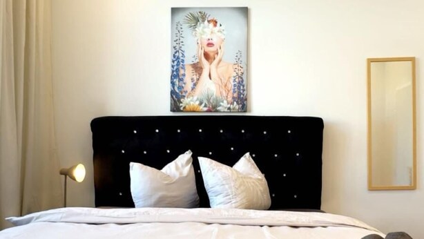 A neatly made bed with white linens and pillows, a black headboard with small decorative elements, a rolled-up dark towel, a framed artwork on the wall, and a floor lamp.