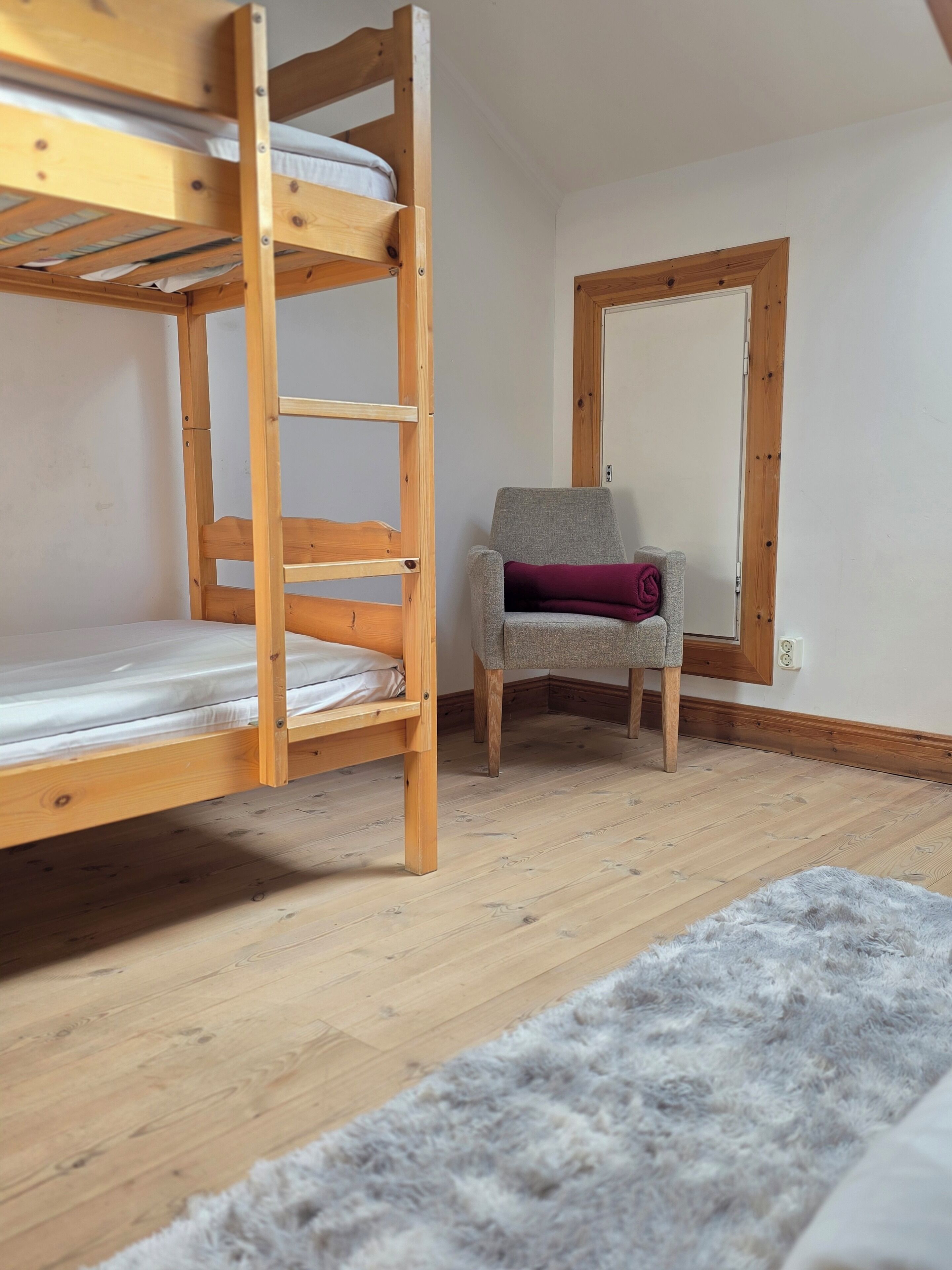 A bunk bed with a ladder, a gray armchair with a purple cushion, a fluffy gray rug, and a wooden-framed window.