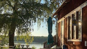 A wooden cabin with a deck overlooking a lake, picnic tables, and a covered umbrella. - Siaröfortet (Ljustero)
