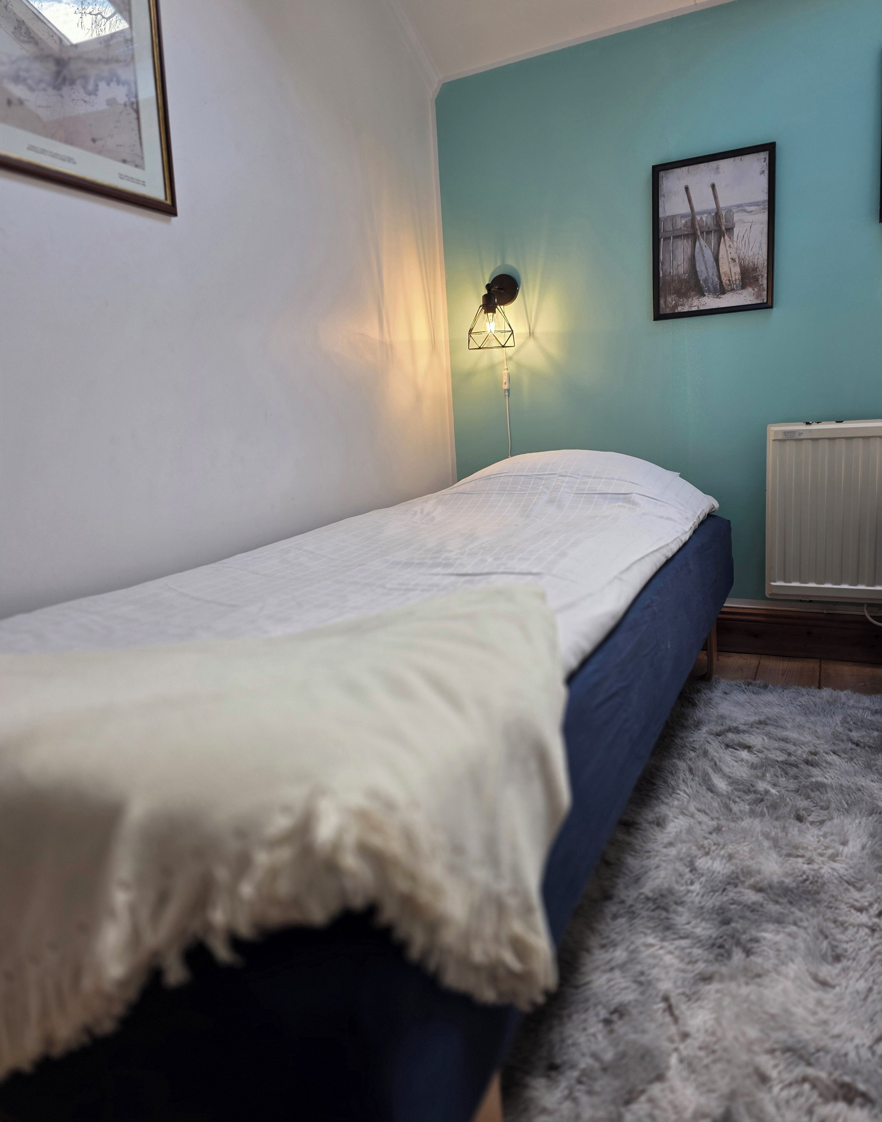 A single bed with a white blanket, a wall-mounted lamp, and a framed picture on a teal wall.