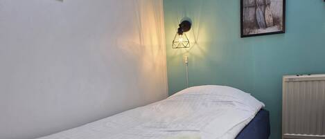 A single bed with a white blanket, a wall-mounted lamp, and a framed picture on a teal wall.