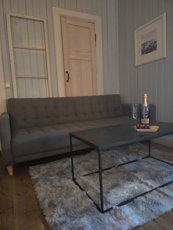 A living room with a grey sofa, a coffee table with a bottle of champagne, glasses, and snacks, a fluffy rug, and a door with a handle. - Siaröfortet (Ljustero)