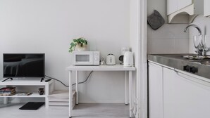 Eine kompakte Küchenzeile mit Mikrowelle, Toaster und Kaffeemaschine auf einer weißen Arbeitsplatte, einem Fernseher auf einem Schrank und einem kleinen Regal mit Büchern und einer Pflanze.