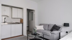 Ein moderner, kompakter Wohnraum mit Küchenzeile, einem grauen Sofa, einem kleinen runden Tisch und einer Nachttischlampe.