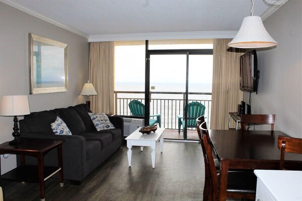 Una sala de estar con sofá, mesa de centro y mesa de comedor. Hay un televisor montado en la pared y un ventanal con vistas al mar.