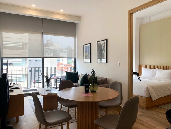 A modern hotel room with a large balcony, a round dining table, and a bed with a wooden headboard.