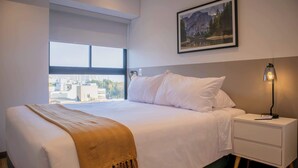 A bedroom with a large bed, a nightstand with a lamp, a window with a city view, and a framed picture on the wall. - w* | Amazing 2BR w/ Balcony in San Isidro (San Isidro)