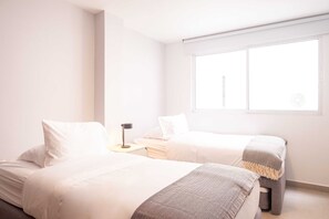 A minimalist bedroom with two beds, a bedside table with a lamp, and a window providing natural light. - w*| Glorious 2BR w/ AC in Bocagrande (Cartagena de Indias)