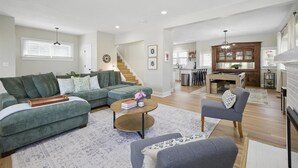 A spacious living room with a sectional sofa, a round coffee table, and a gray armchair. A kitchen with a wooden island and bar stools is visible in the background.