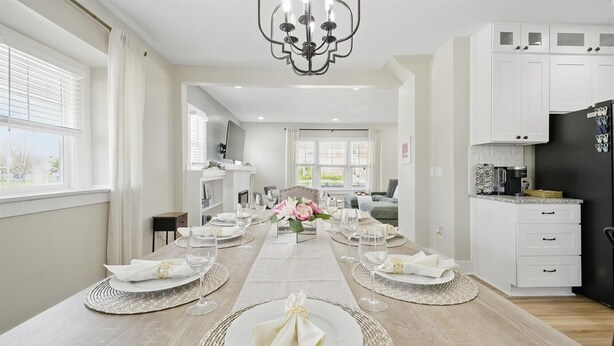 A dining area with a long table set for a meal, a kitchen with white cabinets, and a modern chandelier.
