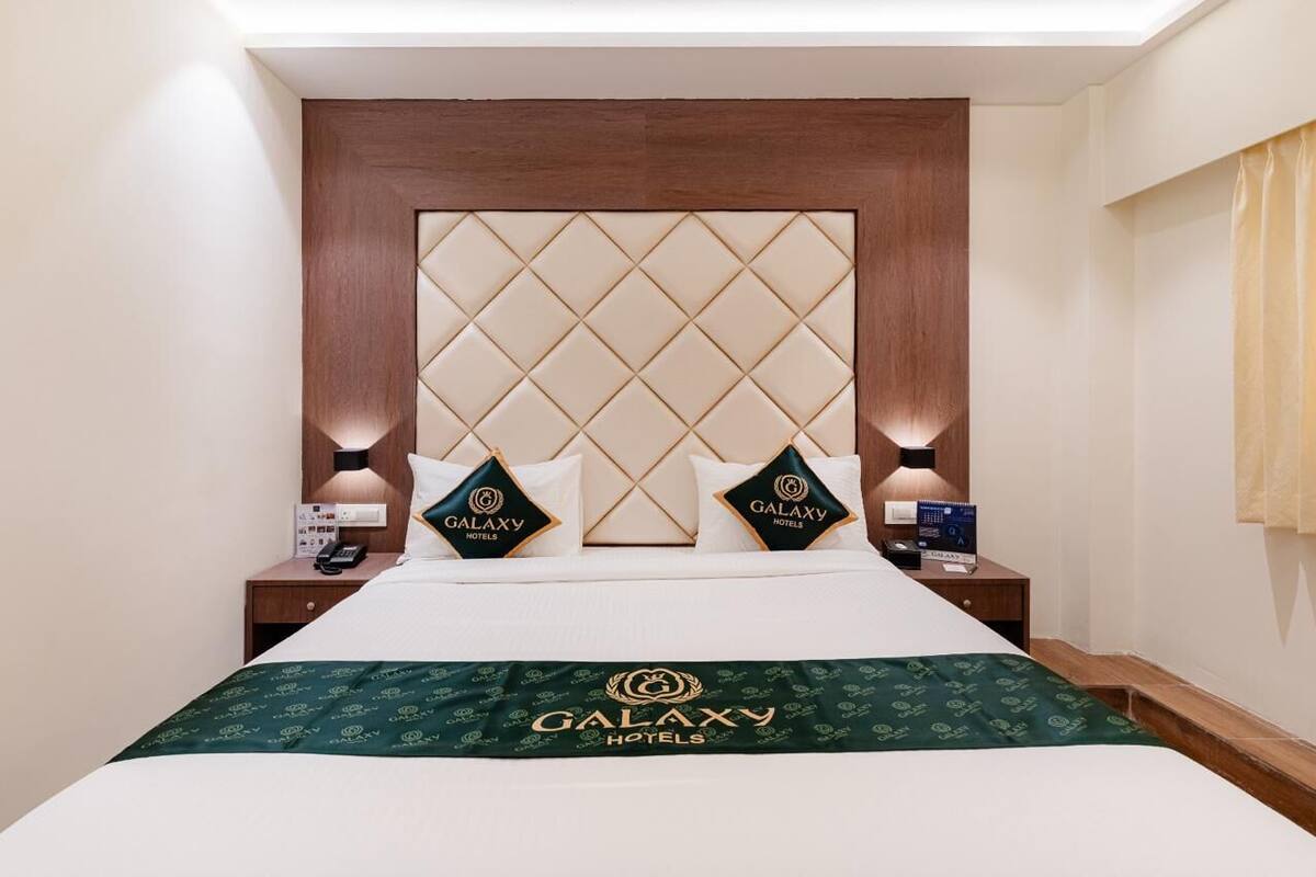 A hotel room with a large bed, a headboard with a diamond pattern, two Galaxy Hotels branded pillows, bedside tables with lamps, and a Galaxy Hotels branded bedspread.