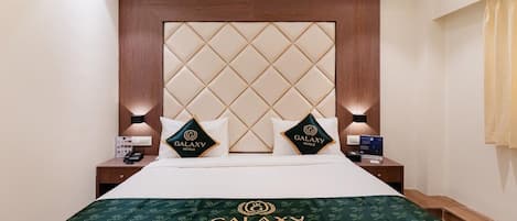 Una habitación de hotel con una cama grande, cabecera con diseño de diamantes, dos almohadas con la marca Galaxy Hotels y mesitas de noche con lámparas, además de una colcha con el logotipo de Galaxy Hotels.
