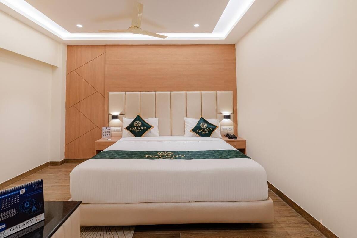 A hotel room with a large bed, a wooden headboard, two bedside tables with lamps, and a Galaxy Hotel logo on the bedspread.