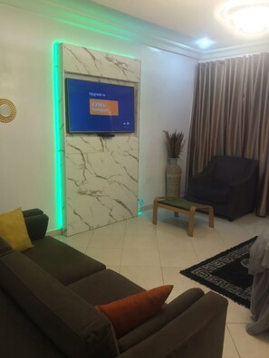 A living room with a sofa, a chair, a small table, and a TV mounted on a marble wall. - Unity Royal Apartment (Lekki)