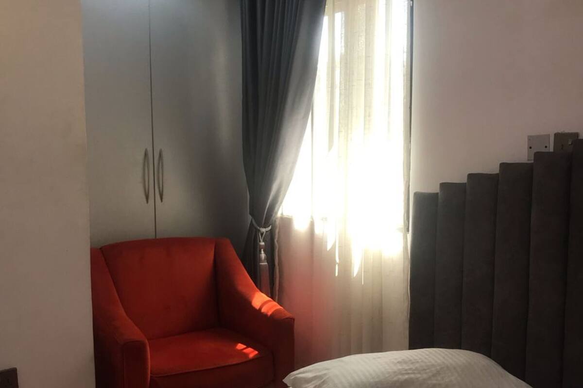 A bedroom with a bed, a red armchair, a wardrobe, and a window with curtains.