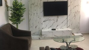 En moderne stue med væg i marmorlook, et fladskærms-tv monteret over et hvidt skab, et glasbord og to sofaer.
