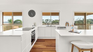 A modern kitchen with white cabinetry, a central island, a clock on the wall, and large windows offering views of the outdoors.