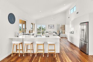 A modern kitchen with a central island, bar stools, a stainless steel refrigerator, and a large clock on the wall. - Corlette Beach Retreat (Corlette)