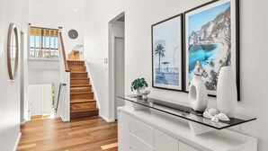 A modern interior with a wooden staircase, a glass console table, and two framed coastal landscape paintings.