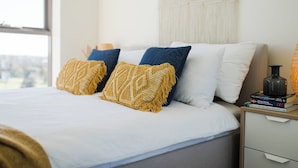 Een net opgemaakt bed met een witte dekbedovertrek en drie decoratieve kussens in geel, blauw en wit.