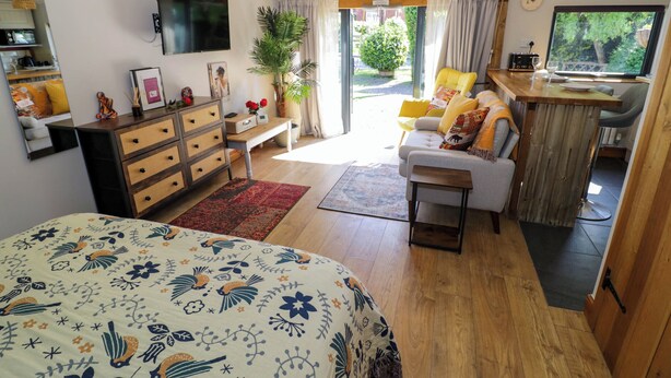 A cozy bedroom with a bed, dresser, and a TV. There is a dining area with a table and chairs, and a large window with curtains. - The Pheasant's Nest (Malvern)