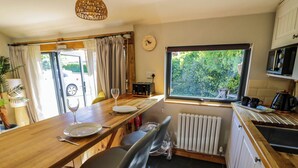 A compact kitchen with a wooden countertop, a dining table with chairs, a clock, and a window with a view of greenery. - The Pheasant's Nest (Malvern)
