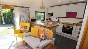 A modern kitchen with white cabinets, a microwave, and an oven. There is a yellow chair with a patterned cushion and a small wooden table. - The Pheasant's Nest (Malvern)