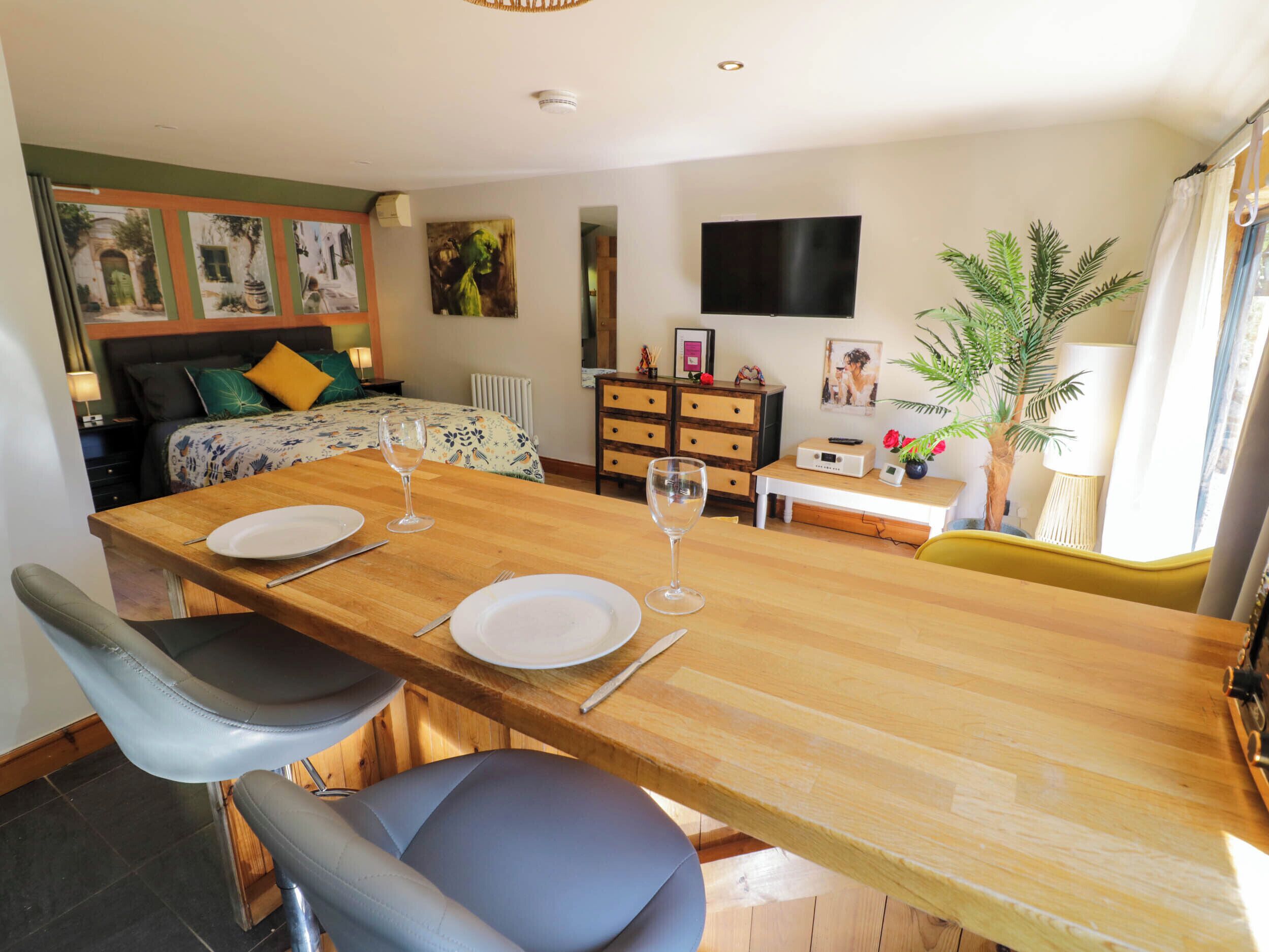 A dining area with a wooden table set for two, a sofa, a TV, and a plant.