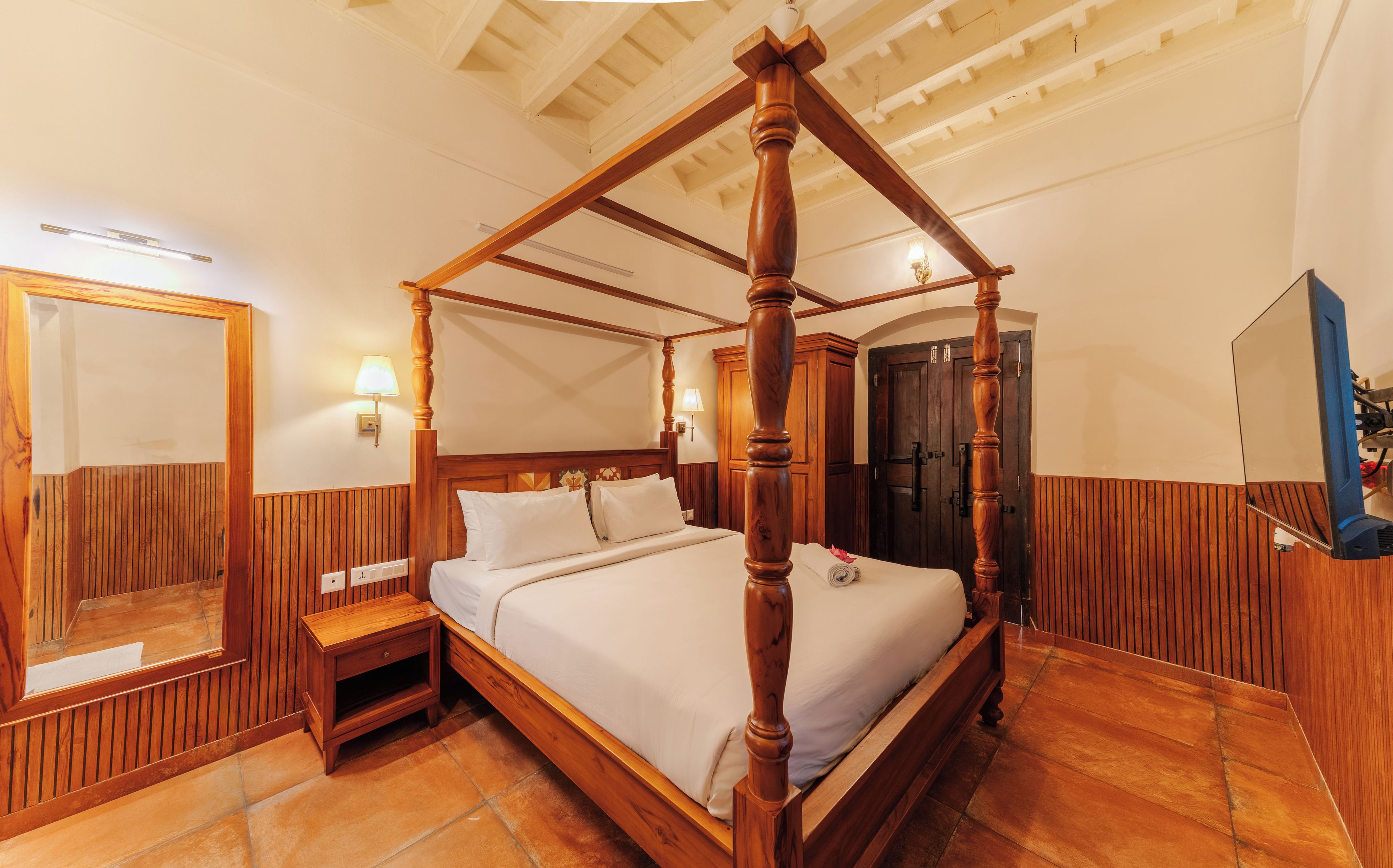 Una cama con dosel de estructura de madera y cabecera, una mesita de noche con espejo y una lámpara fijada a la pared.