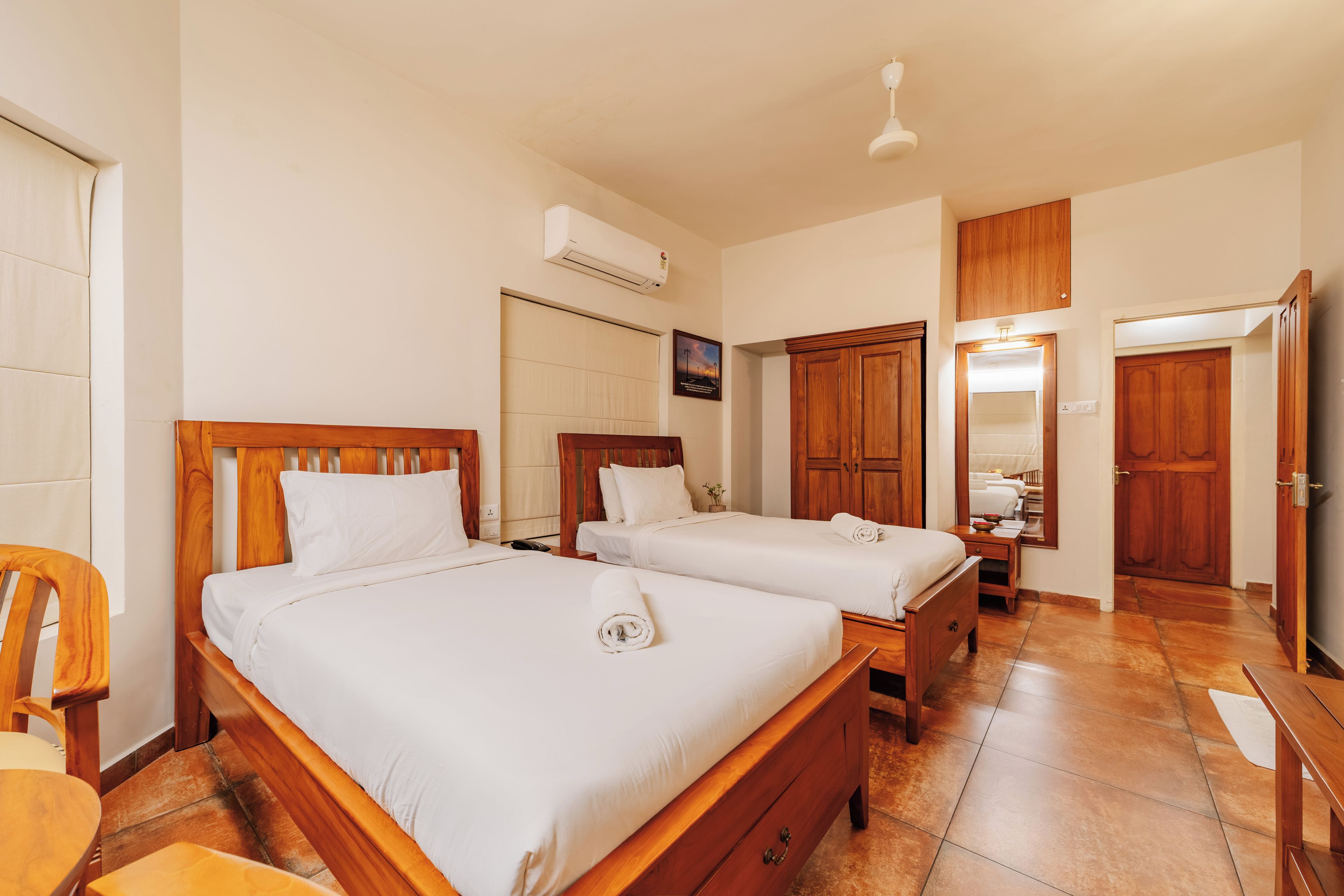 A hotel room with two wooden beds, a wooden wardrobe, a mirror, and a ceiling fan.