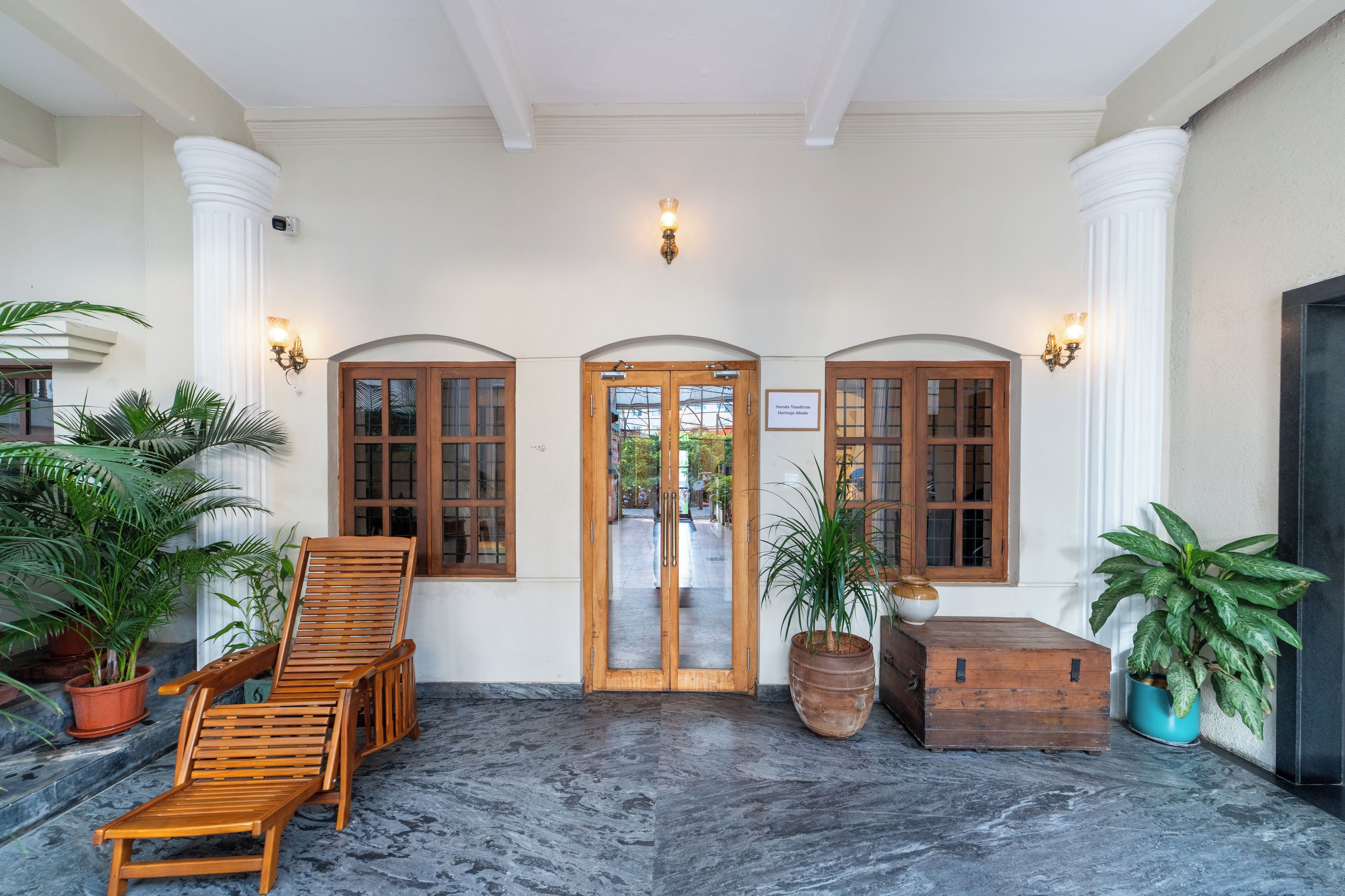 A lobby with a wooden bench, potted plants, and a sign reading 'Sarada Mandiram Heritage Abode'.