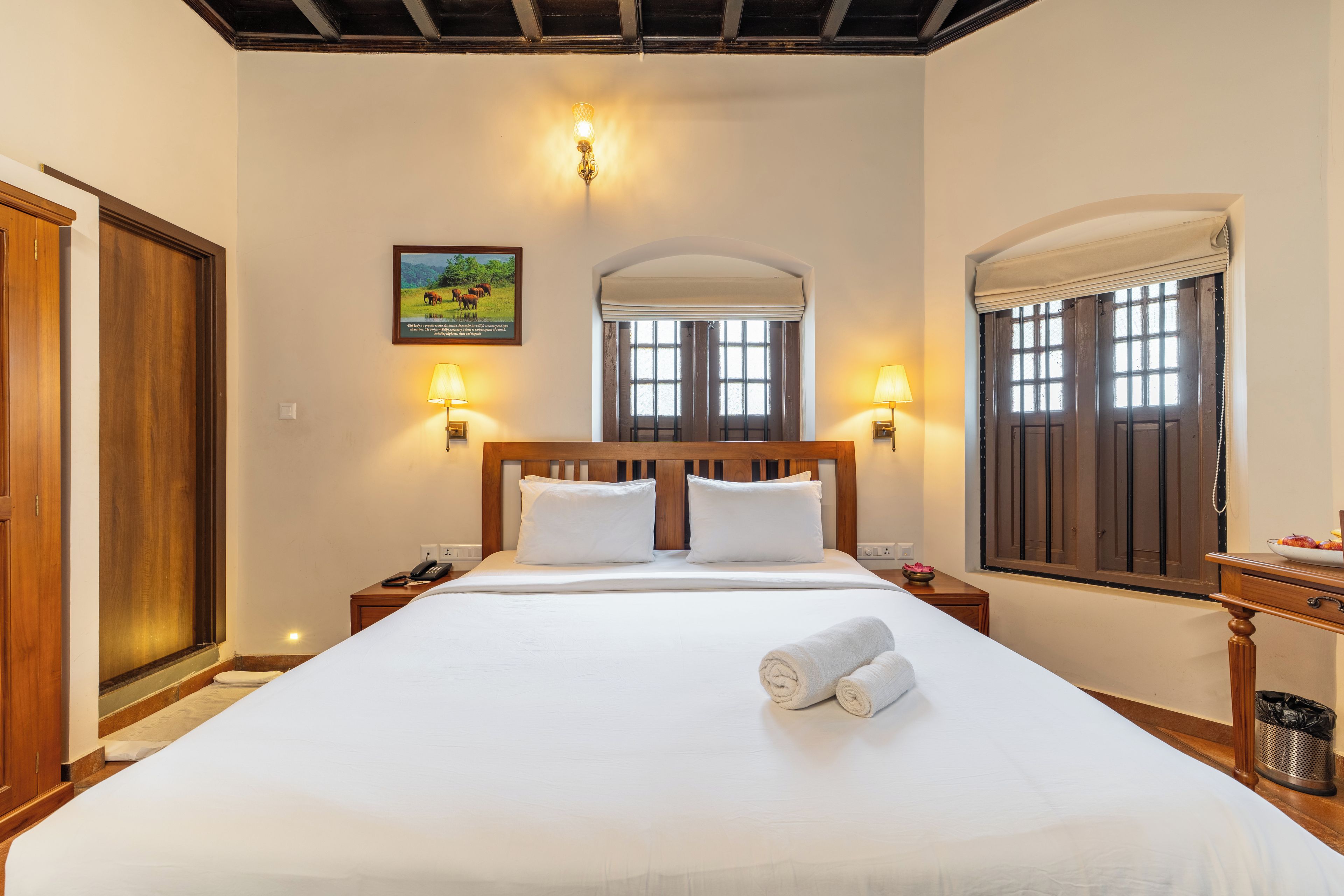 Un cuarto de hotel con una cama grande, cabecera de madera, dos lámparas de noche, un cuadro en la pared y una ventana con vistas al exterior.