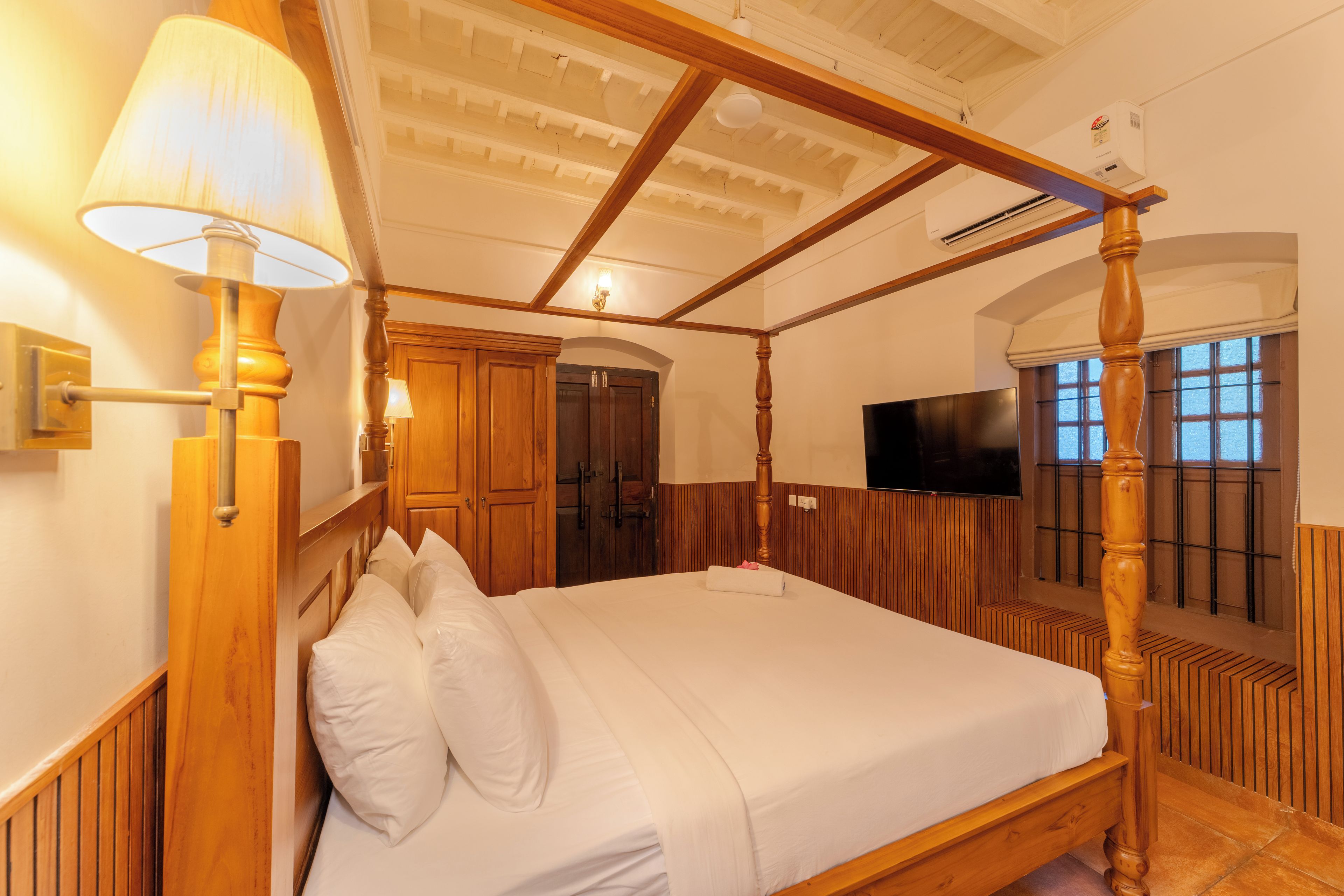 A four-poster bed with white linens, wooden headboard, and a wall-mounted lamp.