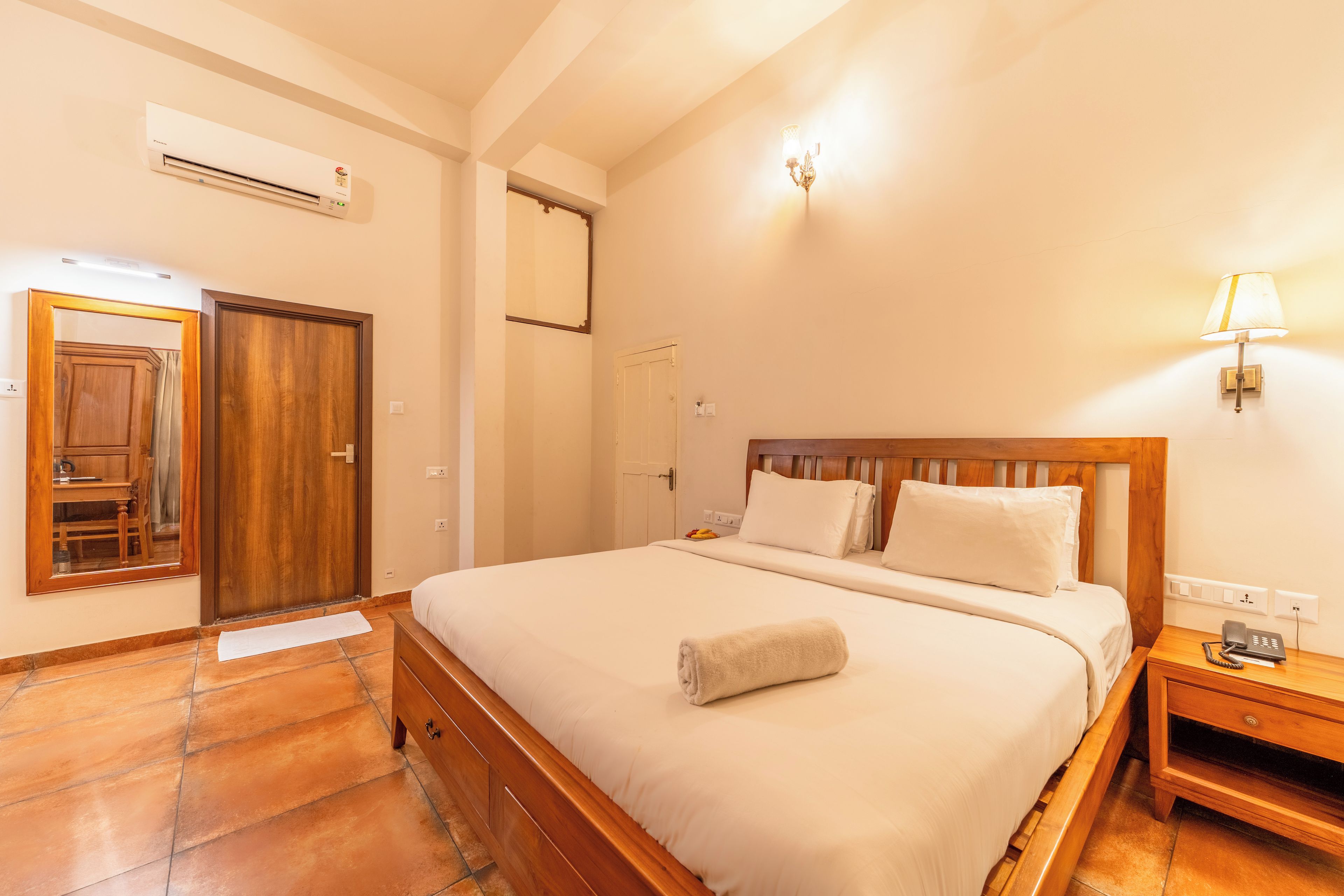 A hotel room with a wooden bed, a bedside table with a lamp, a wardrobe, and an air conditioning unit.